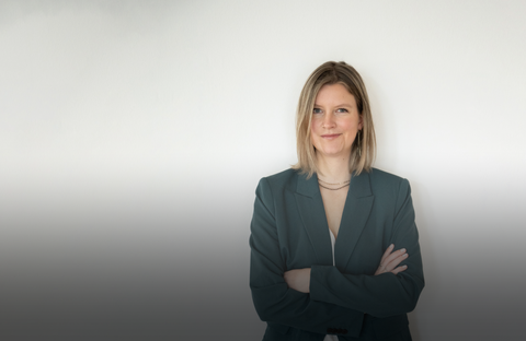 Auf dem Bild ist Sanja zu sehen. Sie trägt einen grünen Blazer, eine weiße Bluse, eine schwarze Leinenhose und schwarze Chucks. Sie hat blonde Haare und eine goldene Kette an. Sie hat die Arme vor der Brust verschränkt und lächelt ganz leicht.