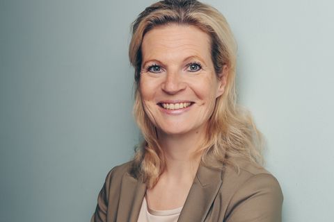 Eine lächelnde Frau mit blonden Haaren in einem beigen Blazer, die die Arme verschränkt hat. - Coaching Komplizin
