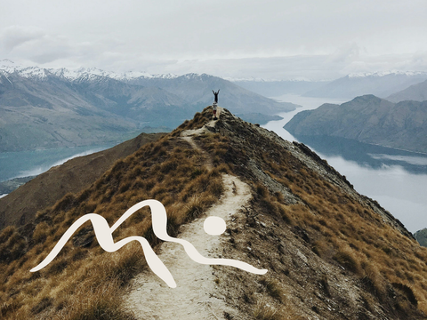 Ein Wanderer steht auf der Spitze eines Berges mit Blick auf einen See und Berge in der Ferne. Links unten sind abstrahierte handgezeichnete Berge zu sehen - Mut Mach Lehre