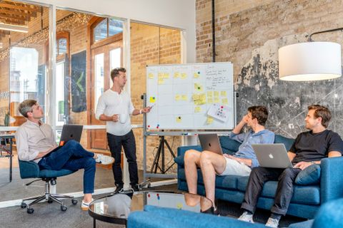 Büroraum mit Whiteboard, Sofas und Laptops, in dem eine Gruppe von Mitarbeitern zusammenarbeitet - Coaching Komplizin