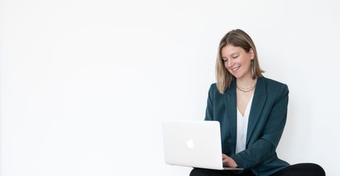 Auf dem Bild ist die Brand Designerin Sanja zu sehen. Sie trägt einen grünen Blazer, eine weiße Bluse, eine schwarze Leinenhose und schwarze Chucks. Sie sitzt auf dem Boden, hat den aufgeklappten Laptop auf den Knien und lächelt. Sanja.Studio | Brand Design