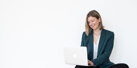 Auf dem Bild ist die Brand Designerin Sanja zu sehen. Sie trägt einen grünen Blazer, eine weiße Bluse, eine schwarze Leinenhose und schwarze Chucks. Sie sitzt auf dem Boden, hat den aufgeklappten Laptop auf den Knien und lächelt. Sanja.Studio | Brand Design