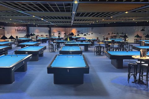 Ein großer Raum mit vielen Billardtischen, die in Reihen angeordnet sind. Der Raum ist dunkel beleuchtet und hat eine moderne, industrielle Atmosphäre.