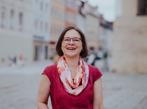 Eine lächelnde Frau mit Brille und rotem Oberteil steht auf einer Straße in einer Stadt. - TANJA BAUER-ROIKE ÜBERSETZT. Deutsch. Englisch. Rechtliches. Offizielles.