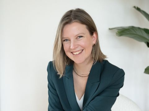 Auf dem Bild ist Sanja zu sehen. Sie trägt einen grünen Blazer, eine weiße Bluse, eine schwarze Leinenhose und schwarze Chucks. Sie sitzt auf eine Sessel, lehnt sich nach vorne und lächelt in die Kamera. Sanja.Studio | Brand Design