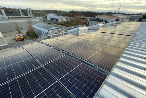 Großes Dach mit vielen Solarpanelen, die Energie aus Sonnenlicht erzeugen. Im Hintergrund sind Industriegebäude und Bäume zu sehen. - Glonn Solar GmbH