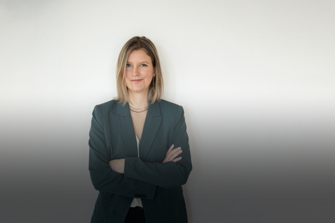 Auf dem Bild ist Sanja zu sehen. Sie trägt einen grünen Blazer, eine weiße Bluse, eine schwarze Leinenhose und schwarze Chucks. Sie hat blonde Haare und eine goldene Kette an. Sie hat die Arme vor der Brust verschränkt und lächelt ganz leicht.