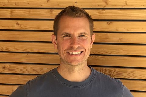 Ein lächelnder Mann mit kurzen blonden Haaren, der vor einer Holzwand steht und eine graue T-Shirt trägt. - QPLAN Engineering