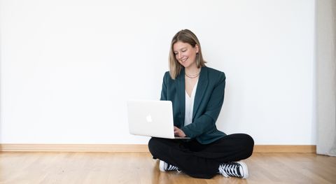Auf dem Bild ist die Brand Designerin Sanja zu sehen. Sie trägt einen grünen Blazer, eine weiße Bluse, eine schwarze Leinenhose und schwarze Chucks. Sie sitzt auf dem Boden, hat den aufgeklappten Laptop auf den Knien und lächelt. Sanja.Studio | Brand Design