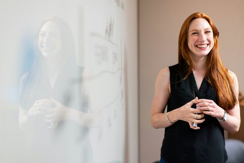 Eine lächelnde Frau mit langen roten Haaren steht vor einer weißen Wand. - Coaching Komplizin