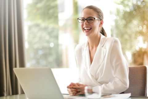 Eine lächelnde Frau in einem weißen Hemd sitzt an einem Laptop und arbeitet in einem hellen Büroraum. - Coaching Komplizin