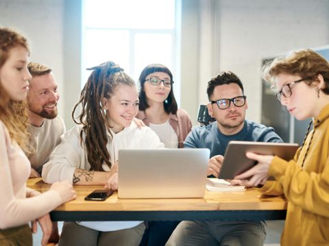 Eine Gruppe junger Menschen, die zusammen an einem Tisch sitzen und an Laptops und Tablets arbeiten. Sie scheinen in einer Besprechung oder Gruppenarbeit vertieft zu sein. - Mut Mach Lehre