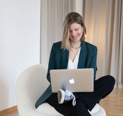 Auf dem Bild ist Sanja zu sehen. Sie trägt einen grünen Blazer, eine weiße Bluse, eine schwarze Leinenhose und schwarze Chucks. Sie sitzt auf einem Sessel, hat ein Bein überschlagen und auf dem Bein ihren Laptop stehen. Sie lächelt und schaut in den Laptop. Sanja.Studio | Brand Design
