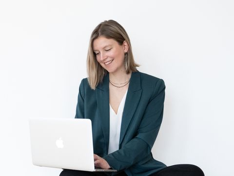 Auf dem Bild ist die Brand Designerin Sanja zu sehen. Sie trägt einen grünen Blazer, eine weiße Bluse, eine schwarze Leinenhose und schwarze Chucks. Sie sitzt auf dem Boden, hat den aufgeklappten Laptop auf den Knien und lächelt. Sanja.Studio | Brand Design