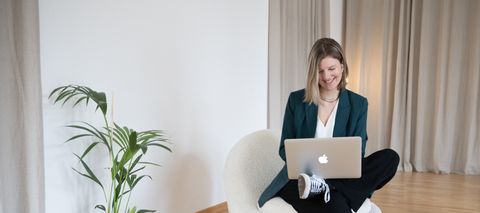 Auf dem Bild ist Sanja zu sehen. Sie trägt einen grünen Blazer, eine weiße Bluse, eine schwarze Leinenhose und schwarze Chucks. Sie sitzt auf einem Sessel, hat ein Bein überschlagen und auf dem Bein ihren Laptop stehen. Sie lächelt und schaut in den Laptop. Sanja.Studio | Brand Design