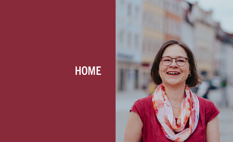 A smiling woman. She wears glasses and a red dress. She stands in front of some buildings. On the left is red text box with 'HOME'.